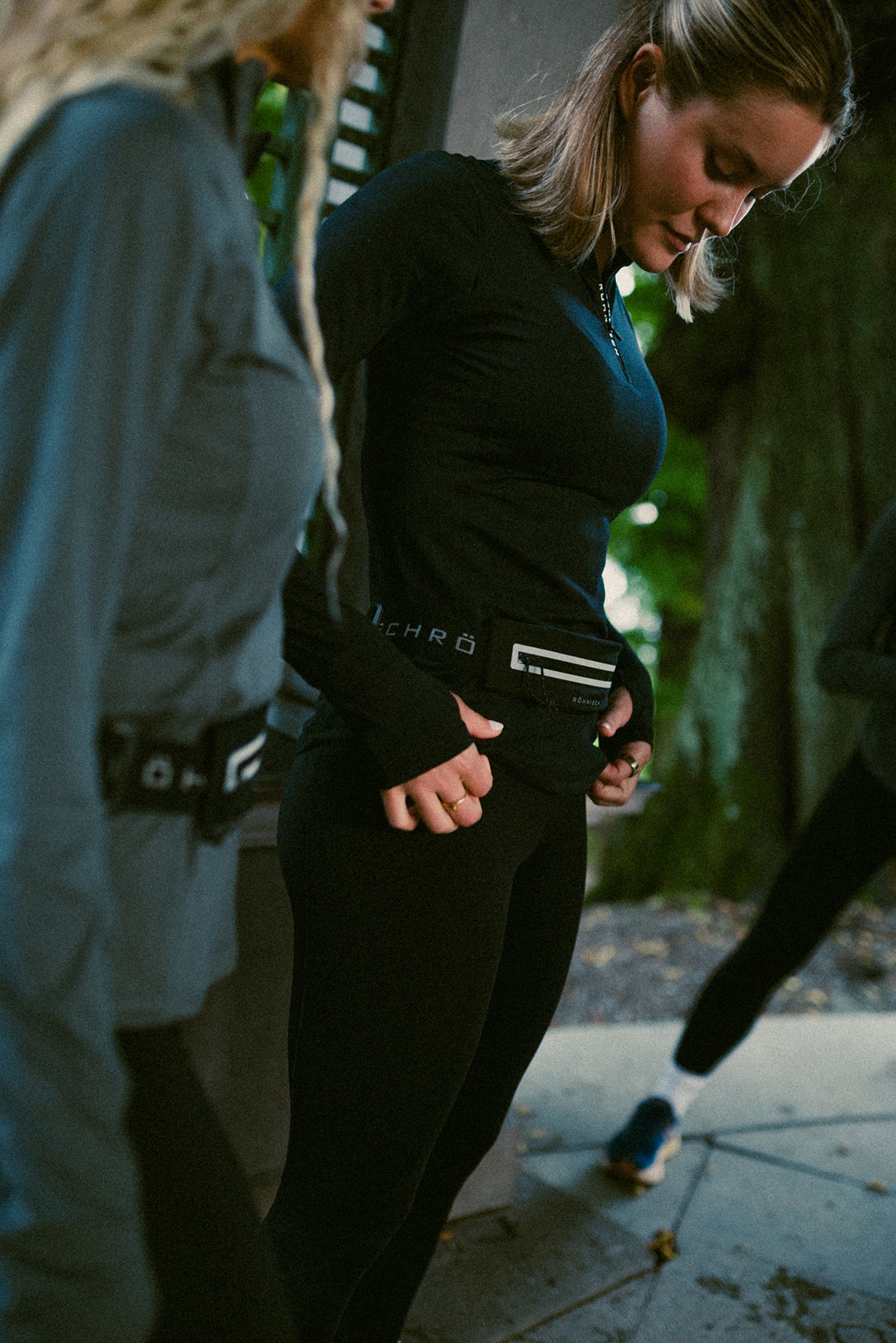 Two women in athletic wear stand outdoors, adjusting waist belts. One looks down, the other is partially visible. Trees and pavement in the background.