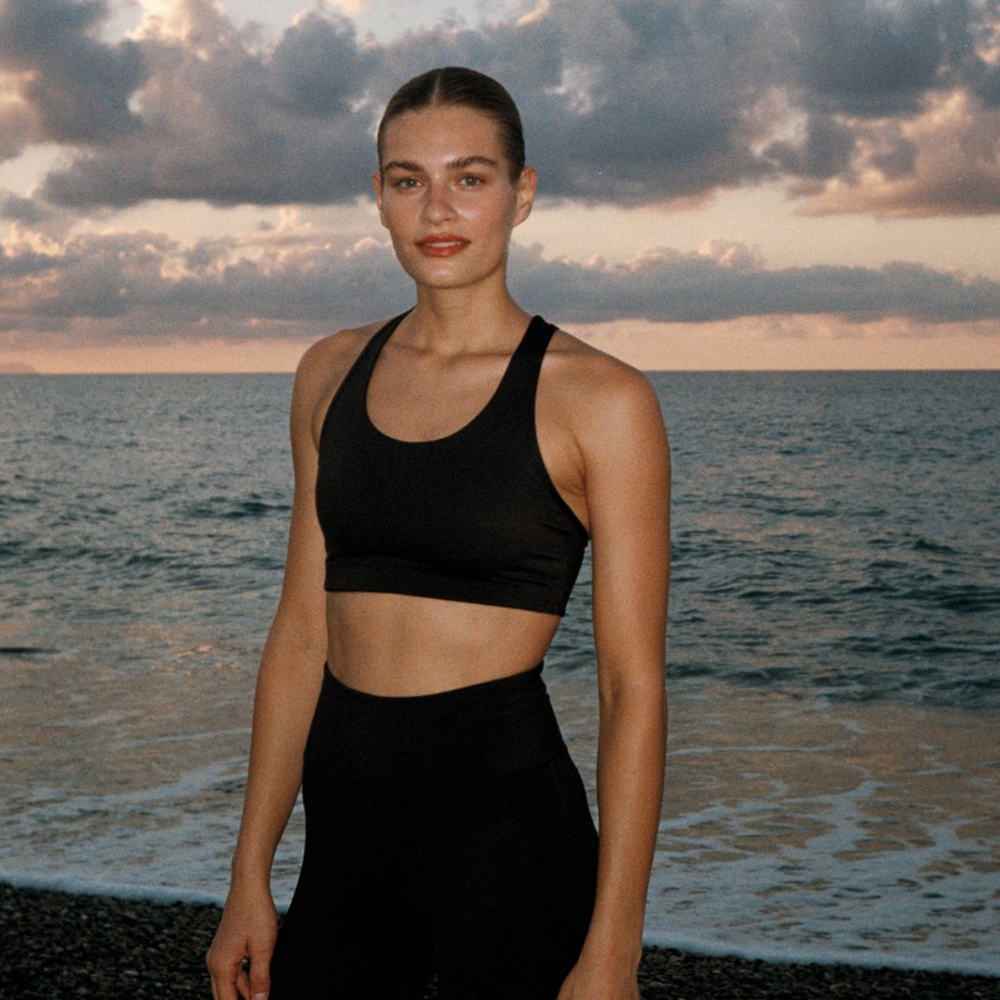 Woman in black workout attire stands on a beach at sunset, with ocean waves and a cloudy sky in the background.