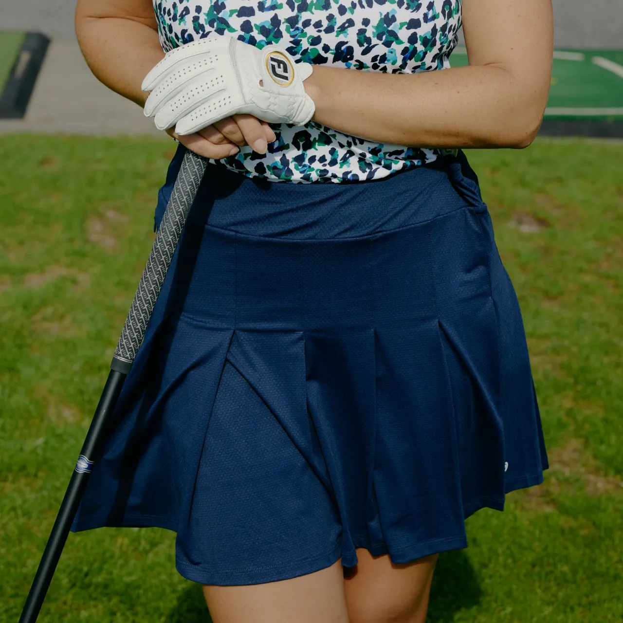 A person wearing a patterned top, navy skirt, and white glove holds a golf club on a grassy area.