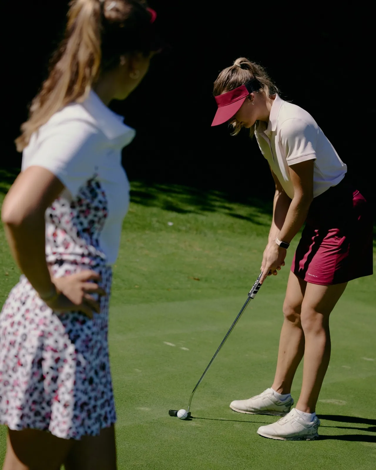 Two women playing golf, one putting the ball while the other watches, on a sunny green course. Both wearing sporty attire and visors.