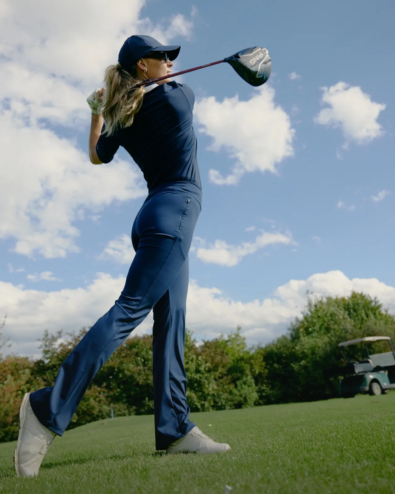 Golfer in blue swinging a club on a grassy course, with trees and a golf cart in the background under a partly cloudy sky.