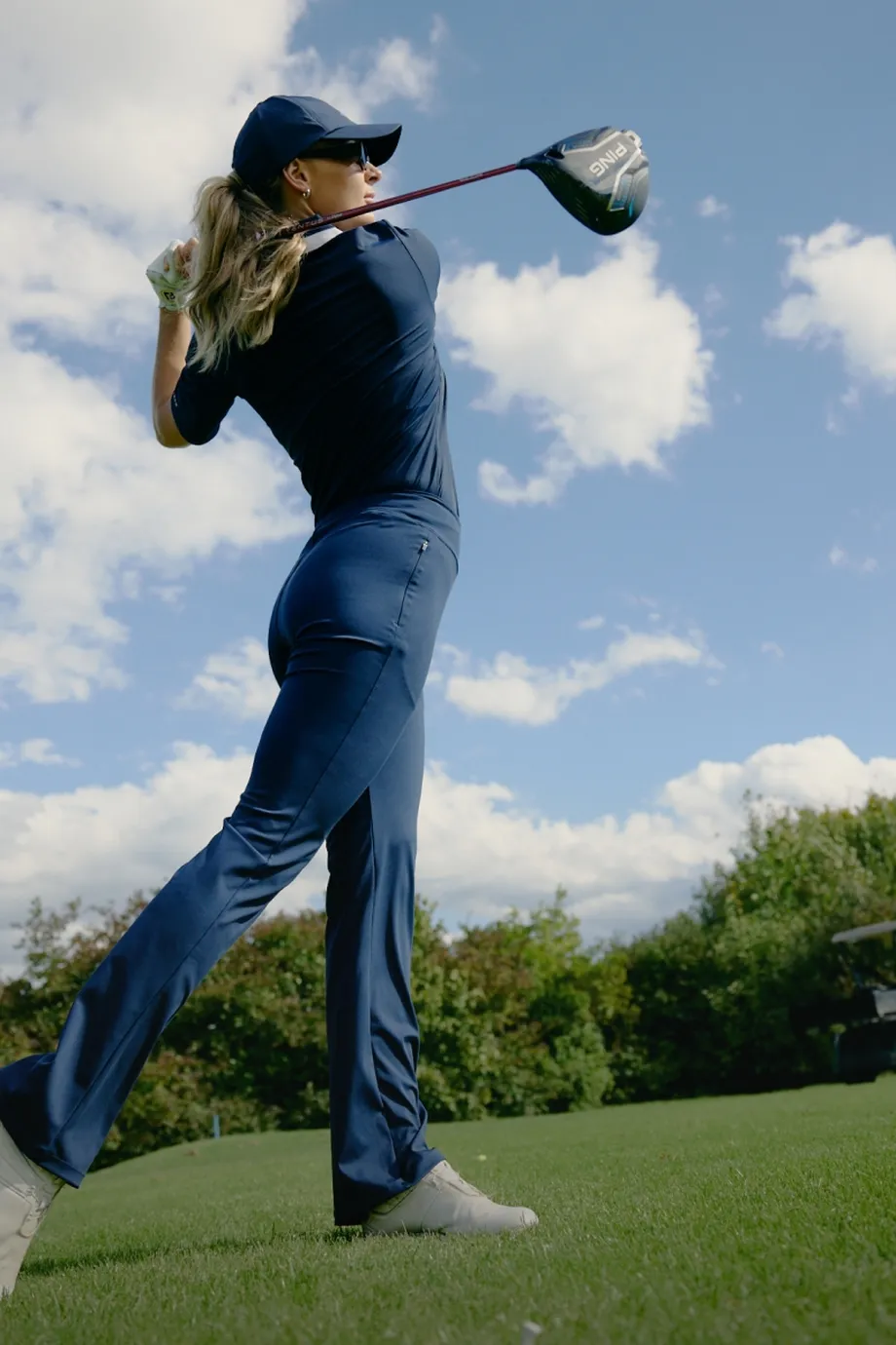 Golfer in blue swinging a club on a grassy course, with trees and a golf cart in the background under a partly cloudy sky.