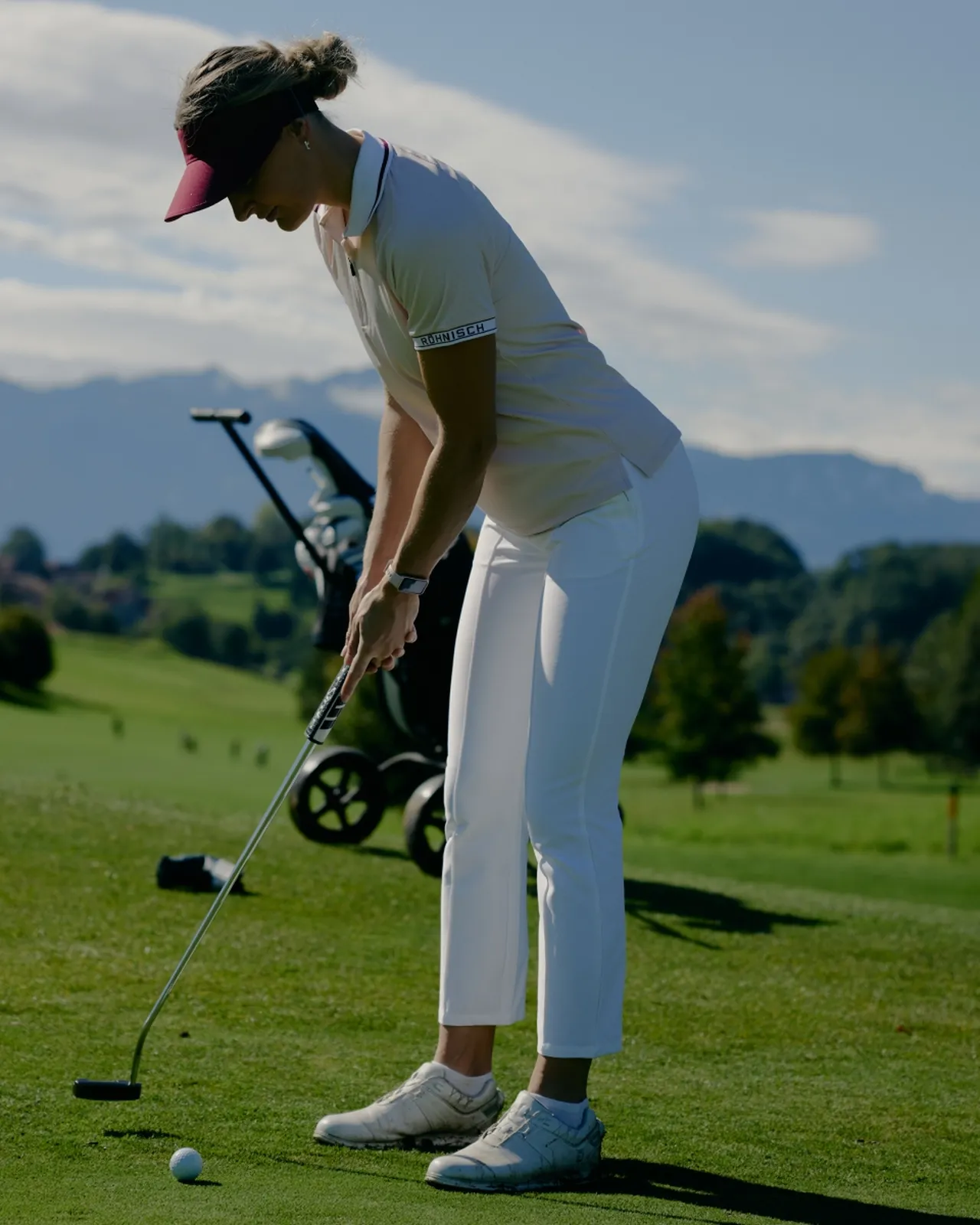 A woman in a visor prepares to putt on a golf course, with a golf cart and scenic mountains in the background on a sunny day.