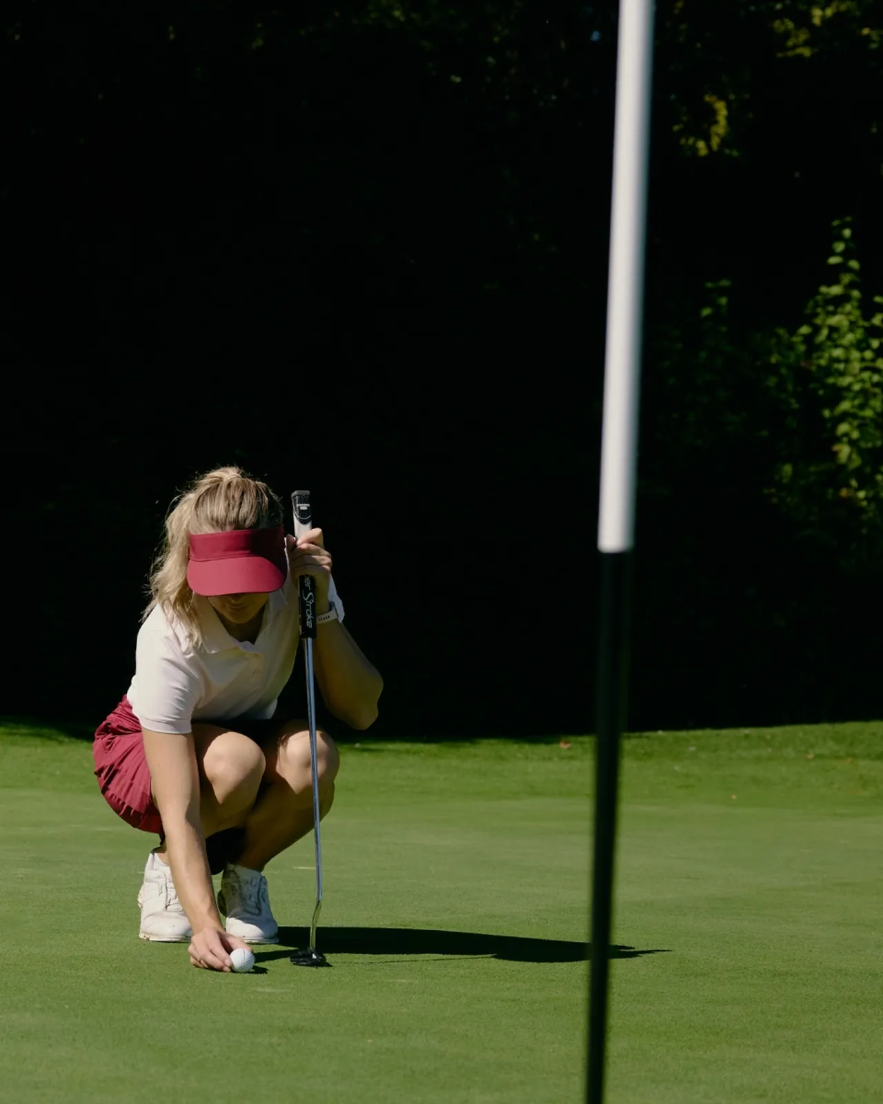 A golfer wearing a red visor and skirt crouches to place a ball on the green near a flagstick, preparing to putt under sunlight.