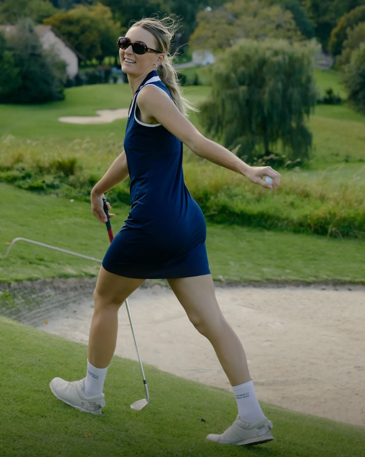 Woman in a blue dress and sunglasses joyfully walking on a golf course, holding a ball and club, with trees and a sand bunker in the background.