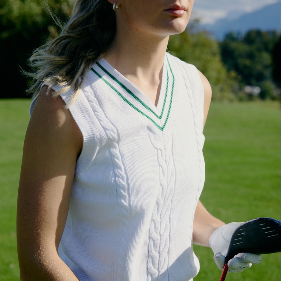 A person in a white sleeveless sweater with green trim holds a golf club on a sunny day, standing in a green field.