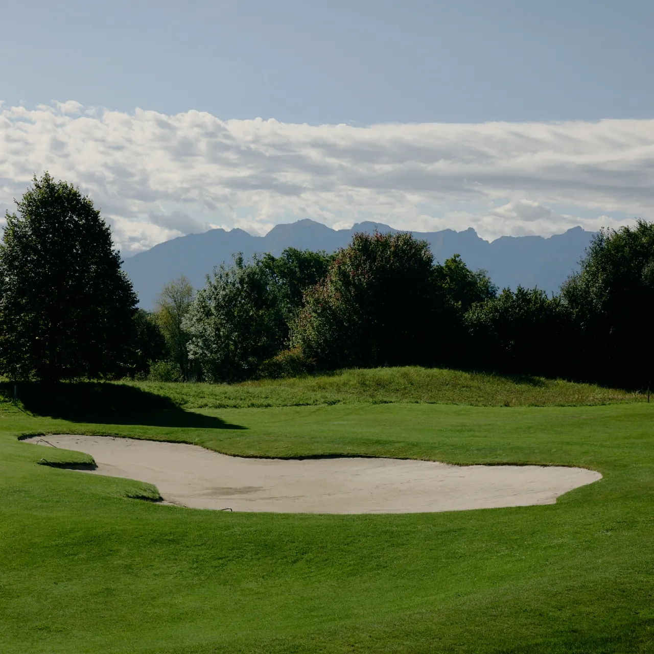 A golf course with a sand trap, lush green grass, trees, and distant mountains under a partly cloudy sky.