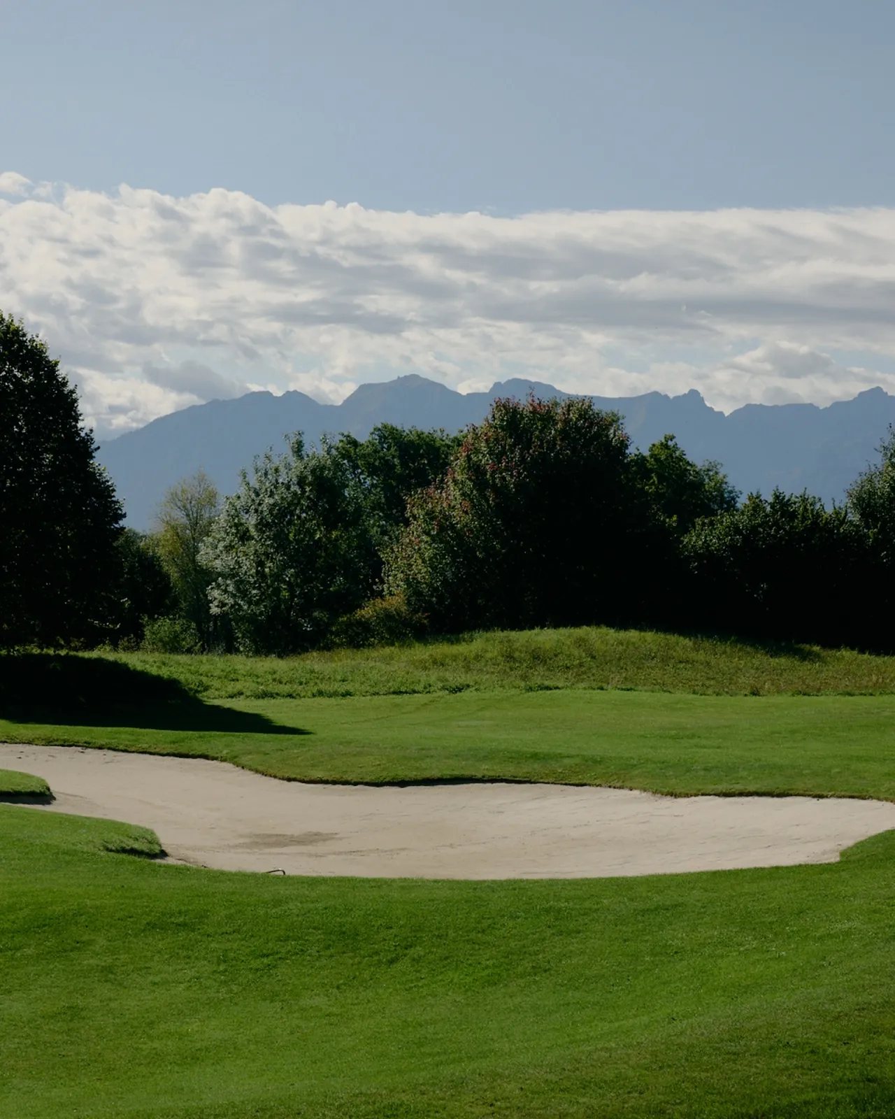 A golf course with a sand trap, lush green grass, trees, and distant mountains under a partly cloudy sky.