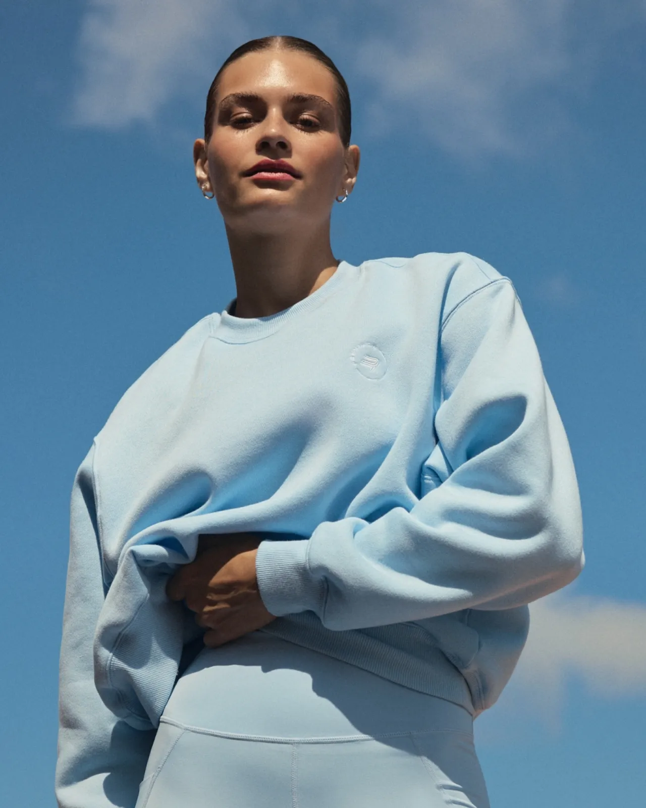 Person in a light blue sweatshirt and leggings stands outdoors under a blue sky with scattered clouds.