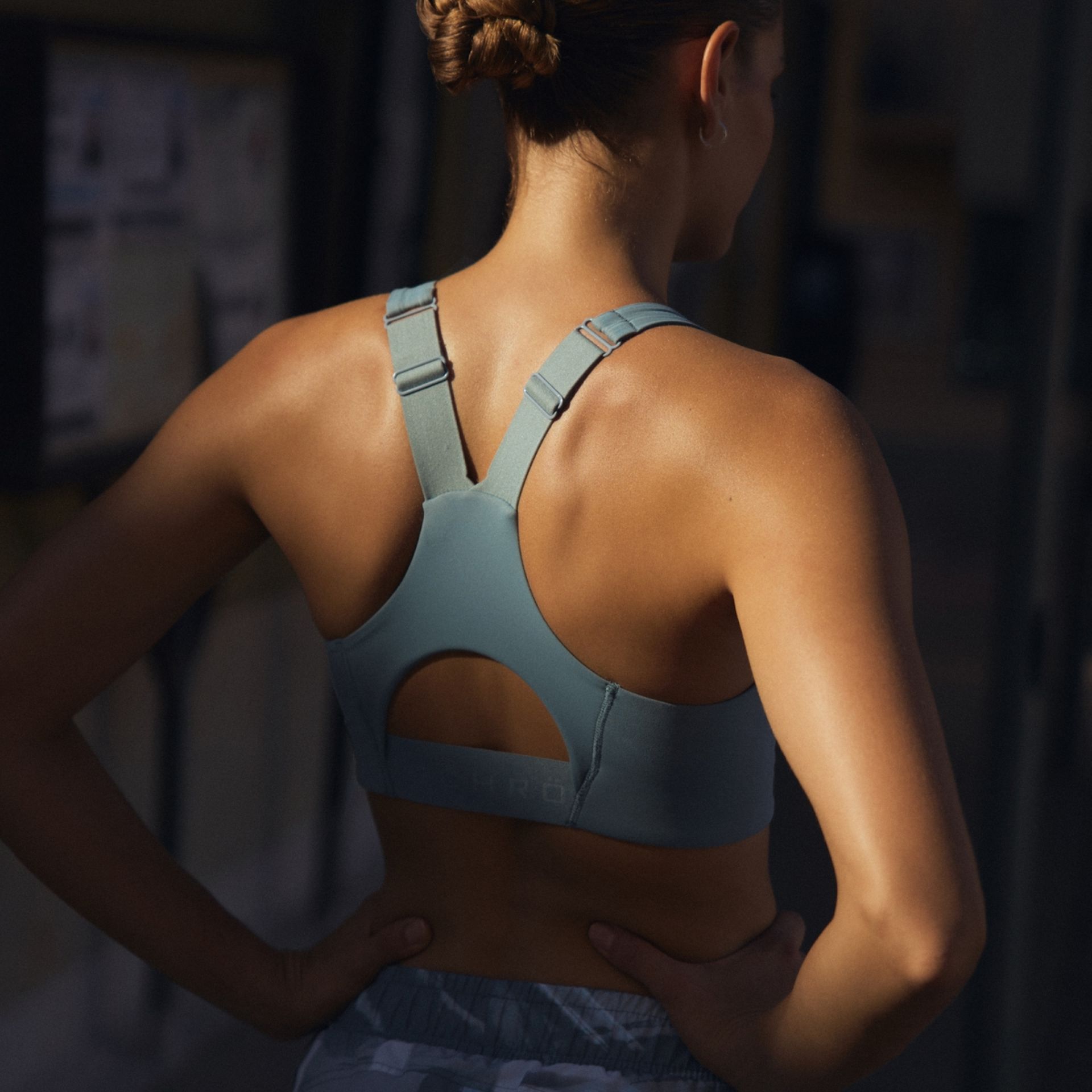 Woman wearing a teal sports bra with adjustable straps, standing outdoors. Her hair is in a bun, and she appears ready for a workout.