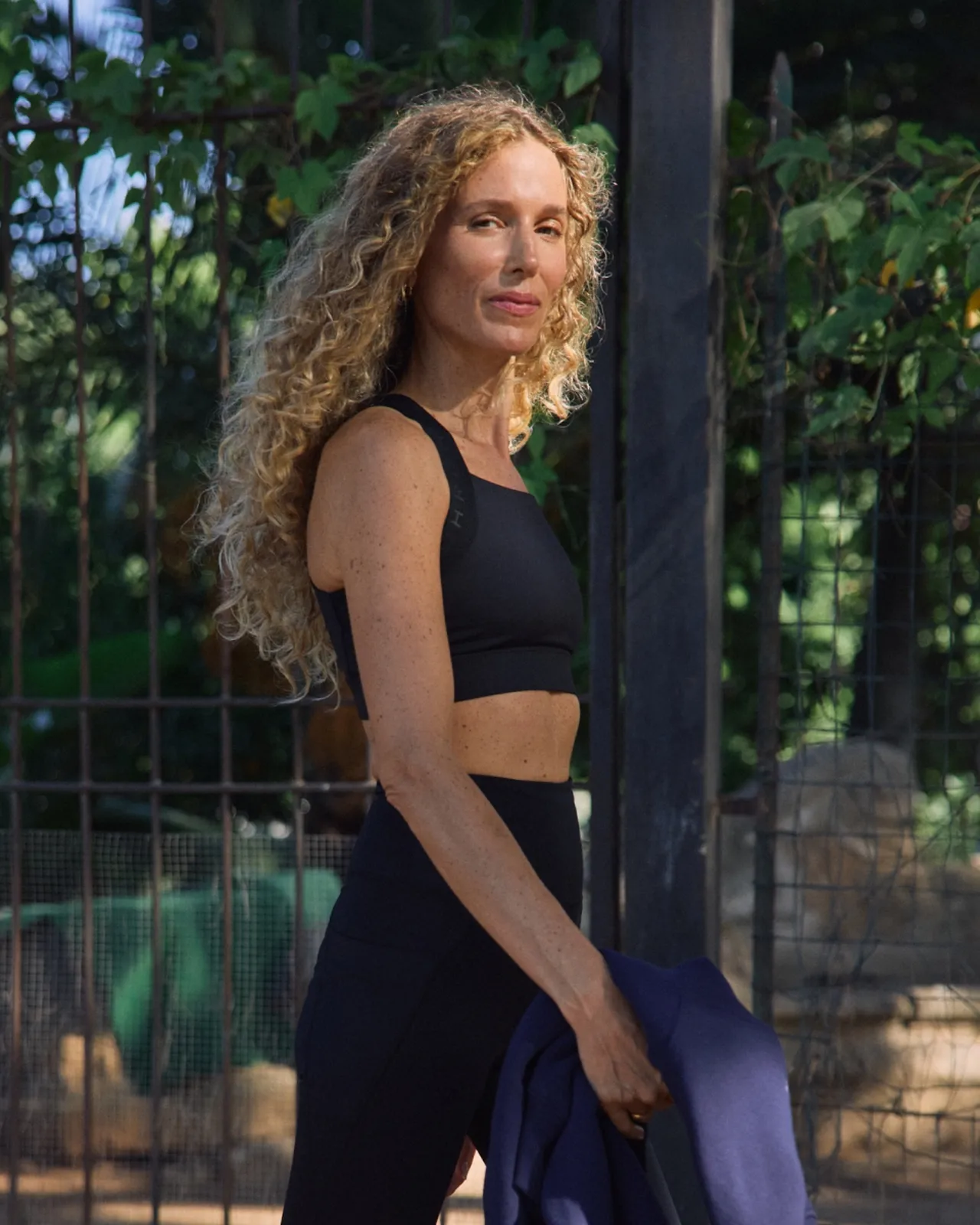 A woman with long curly hair in a black workout outfit holds a jacket, standing outdoors by a fence and lush greenery.
