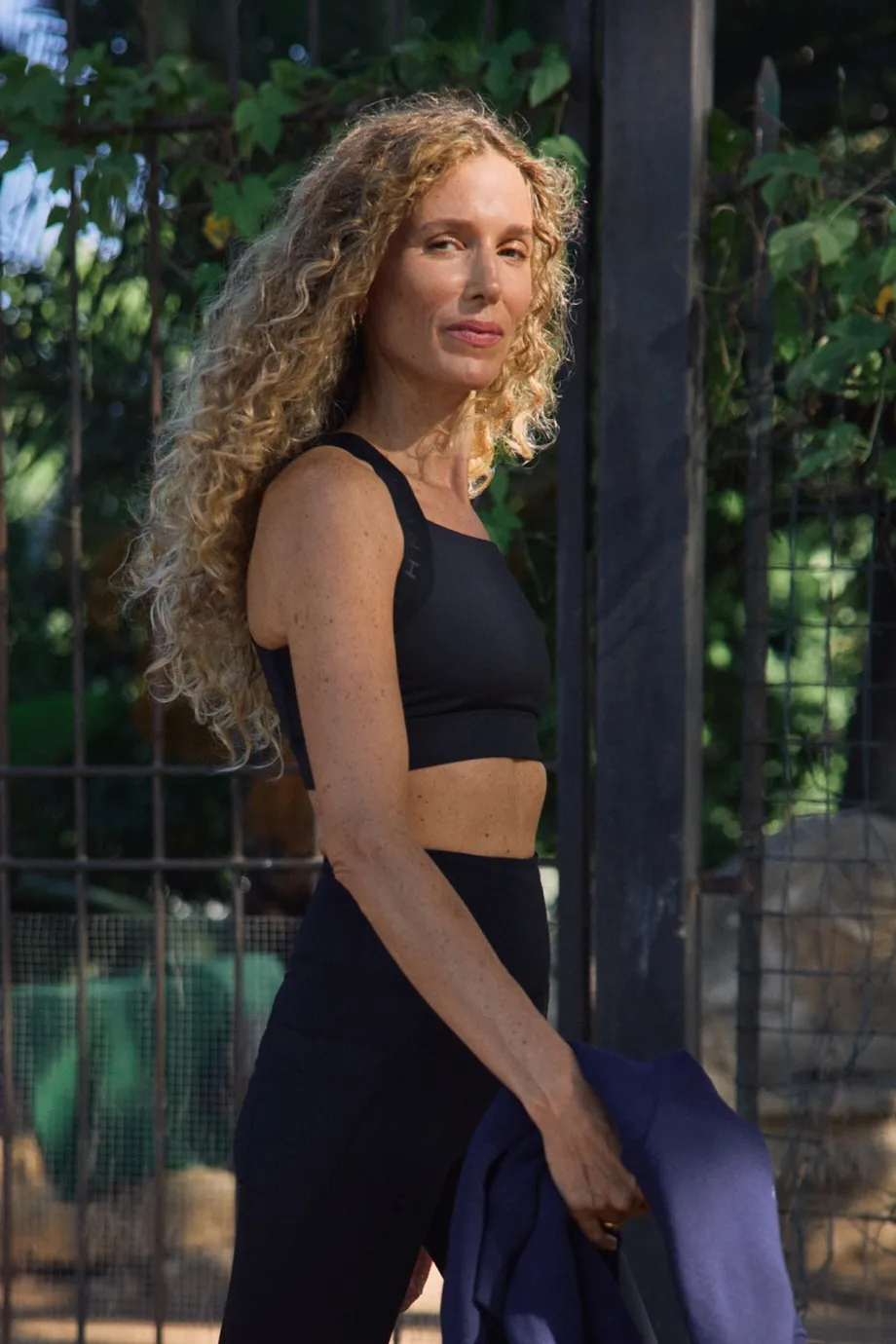A woman with long curly hair in a black workout outfit holds a jacket, standing outdoors by a fence and lush greenery.