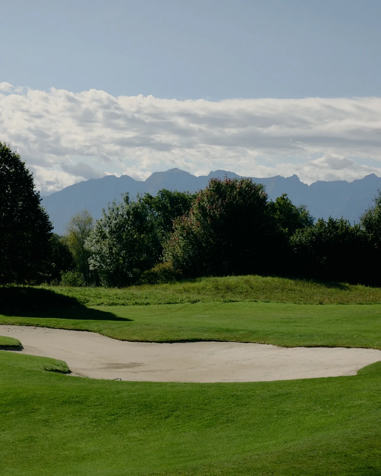 A golf course bunker with lush green grass, surrounded by trees and mountains under a cloudy sky.