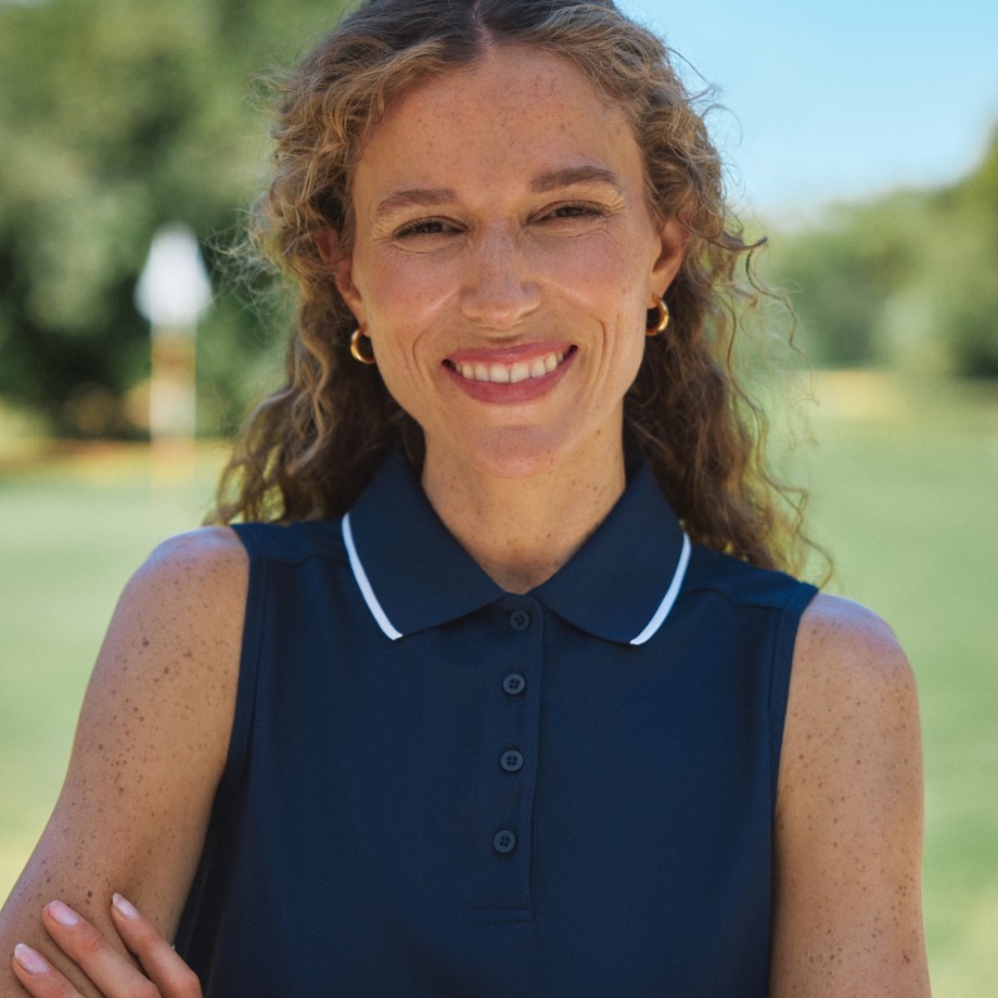 A smiling woman with curly hair and freckles stands outdoors wearing a sleeveless navy polo shirt, with greenery in the background.