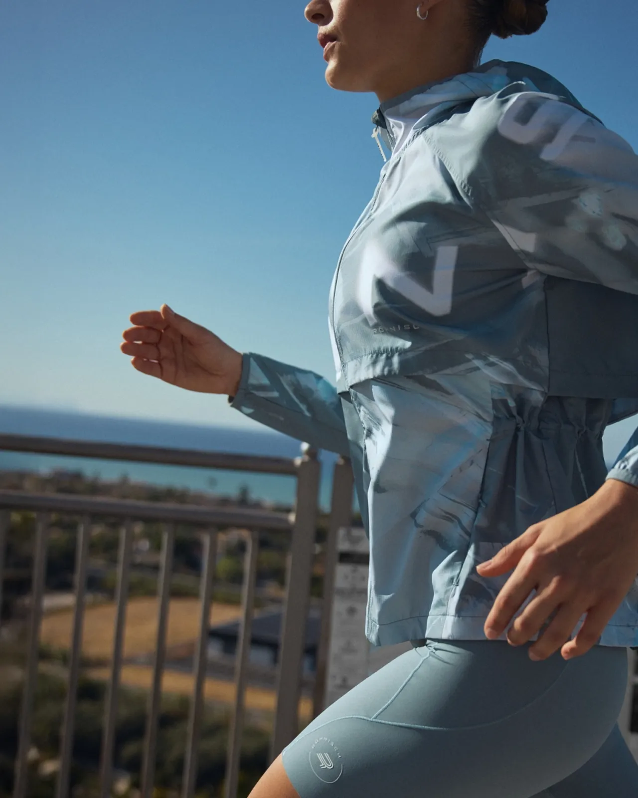 Person in blue activewear jogging outdoors near a railing with the ocean and trees in the background, under a clear blue sky.