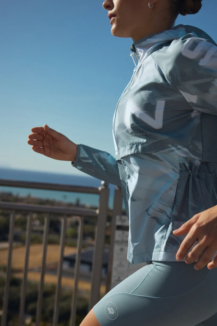 Person in blue activewear jogging outdoors near a railing with the ocean and trees in the background, under a clear blue sky.