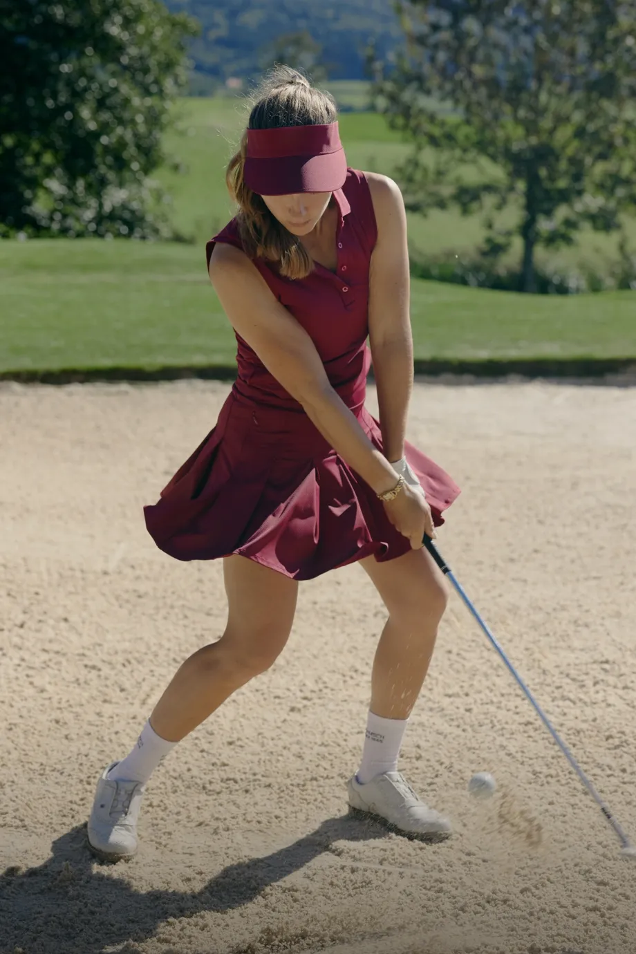 A woman in a maroon golf outfit swings a club in a sand bunker, with grass and trees in the background on a sunny day.