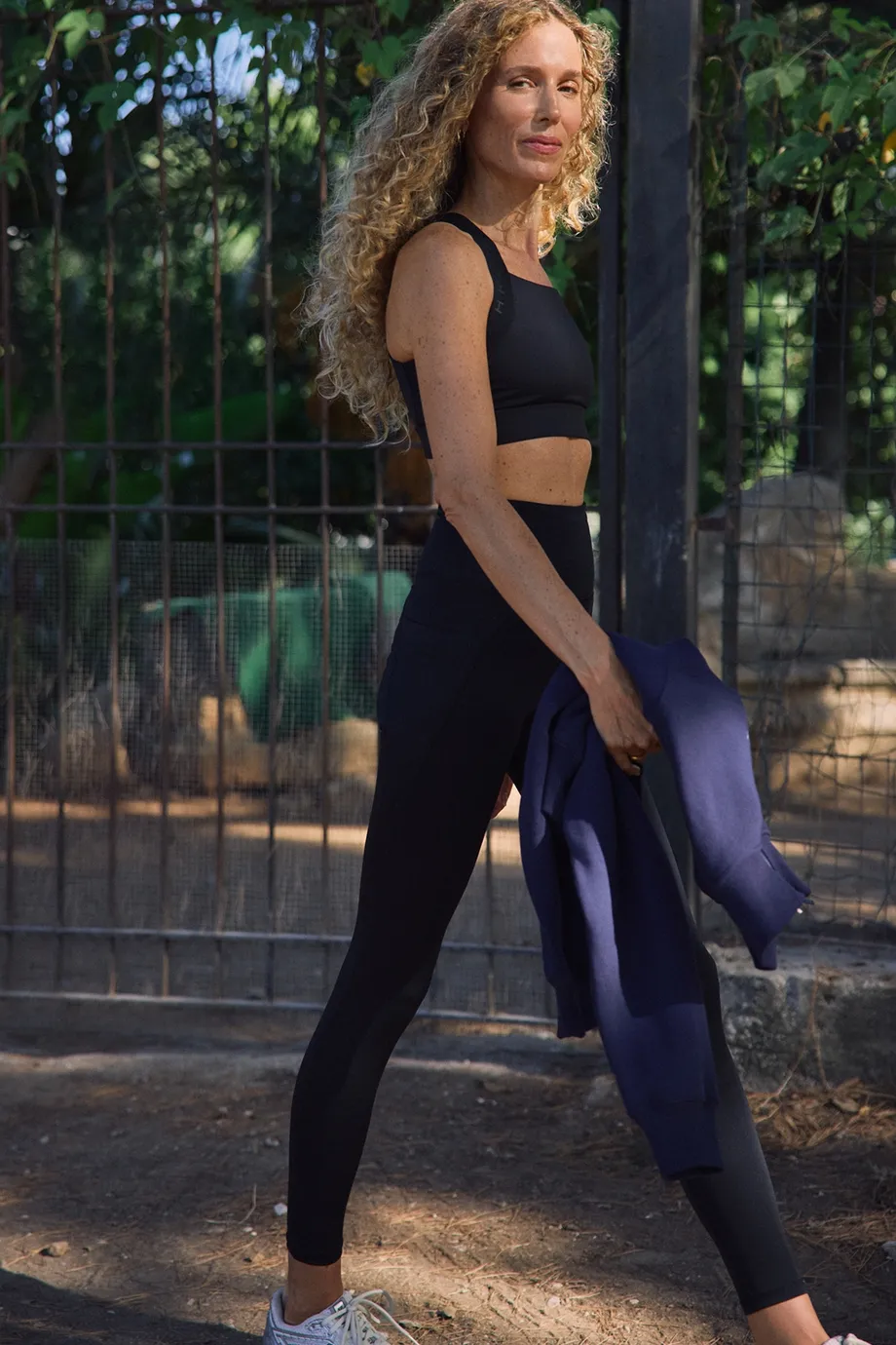 Woman in black athletic wear walks outside, holding a blue jacket. Background includes a fence and greenery.