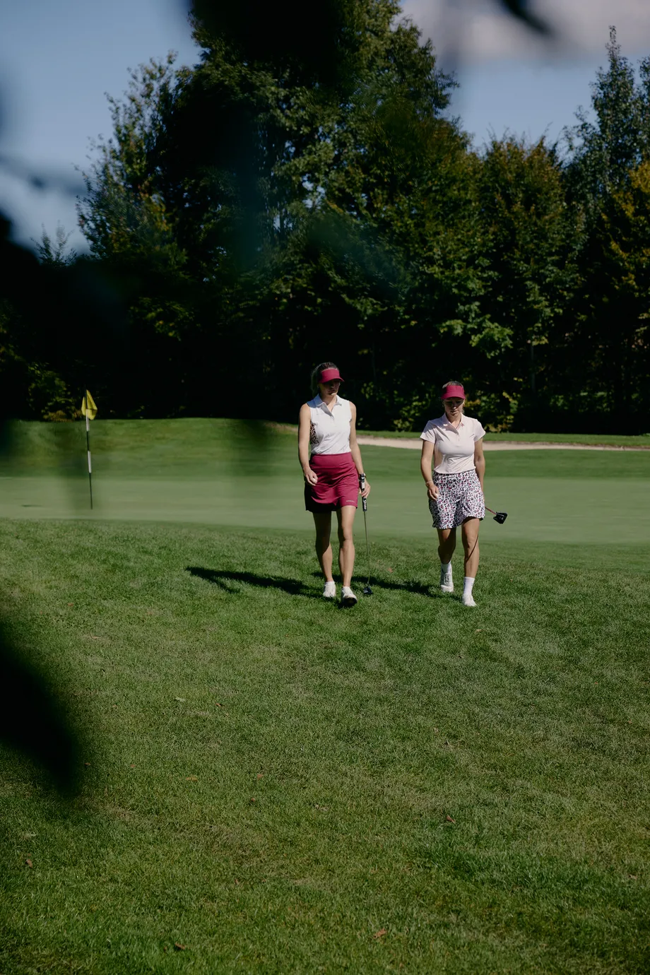 Two women in golf attire walking on a lush green golf course, with trees in the background and a flag in the distance.