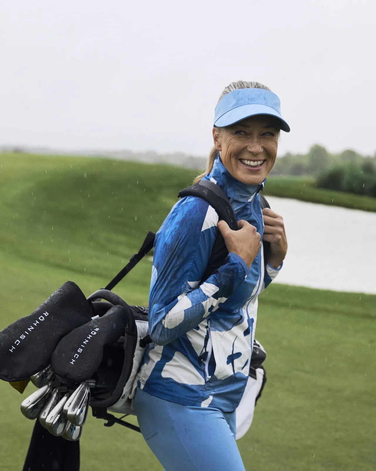 A woman in blue sports attire smiles while carrying a golf bag on a rainy golf course.