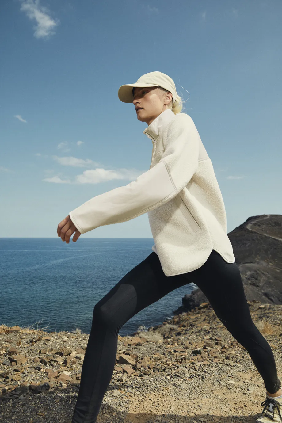 Woman hiking by the ocean in a beige pile jacket, black tights and beige cap