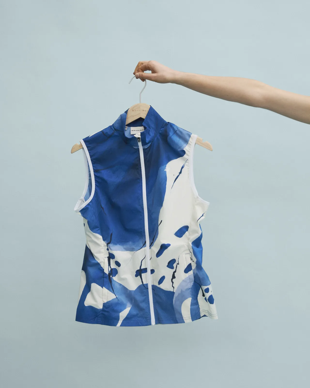 A hand holds a hanger with a sleeveless blue and white abstract-patterned vest against a light blue background.
