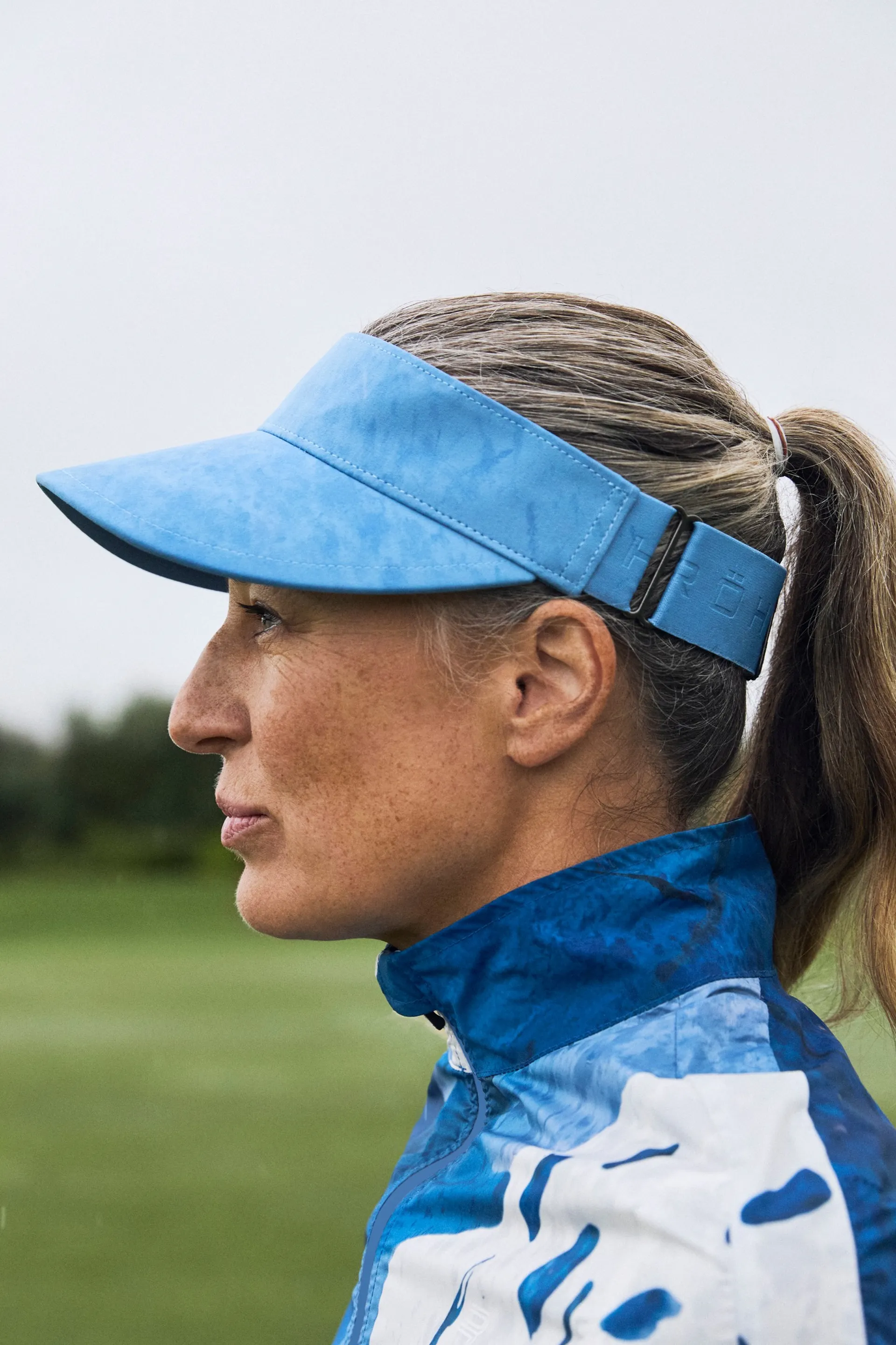 Woman in a blue visor and jacket stands outdoors, facing left. She has a ponytail and is in a grassy area with a cloudy sky.