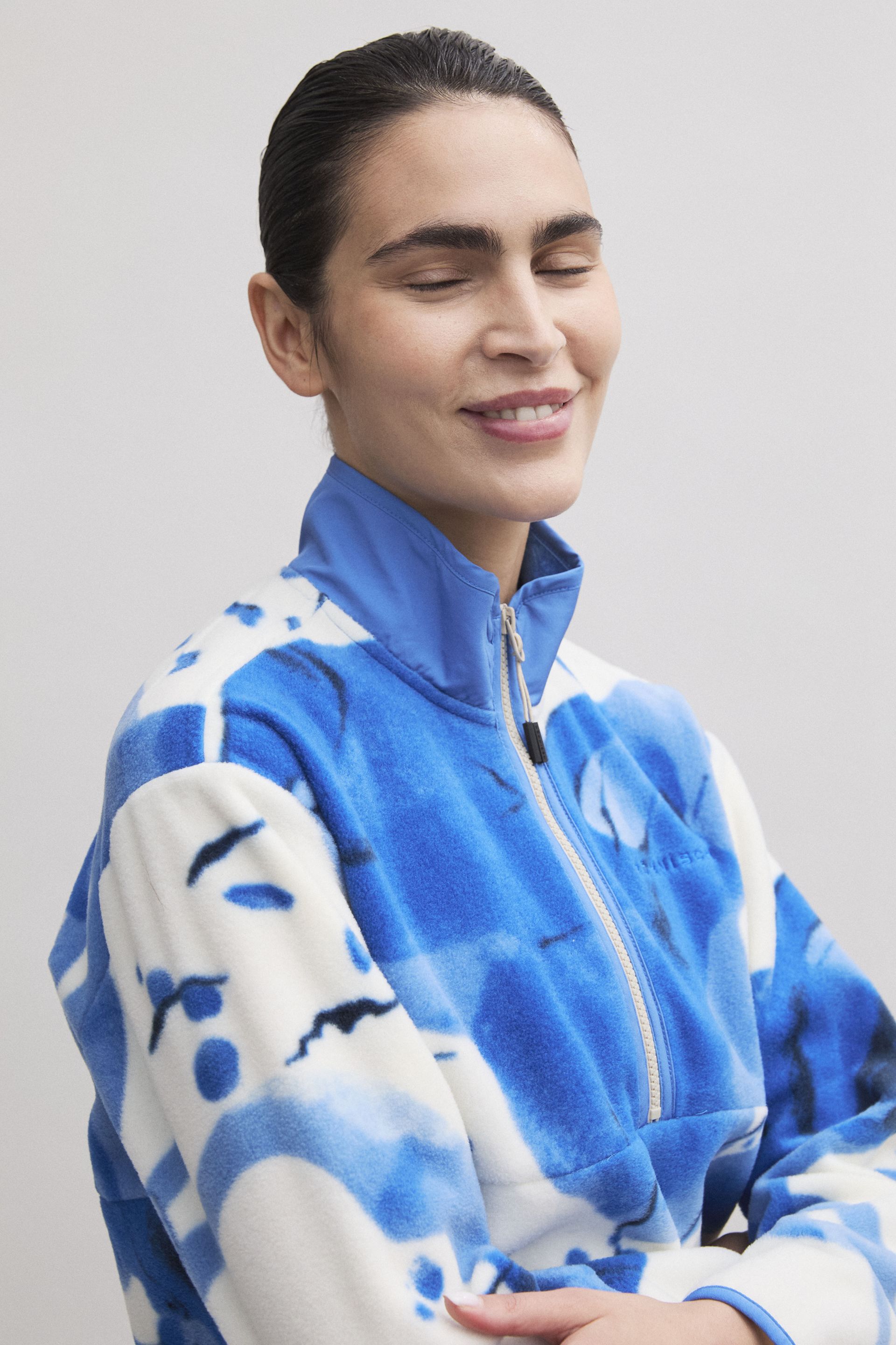A person smiling with closed eyes, wearing a blue and white patterned fleece jacket with a high collar, against a plain background.
