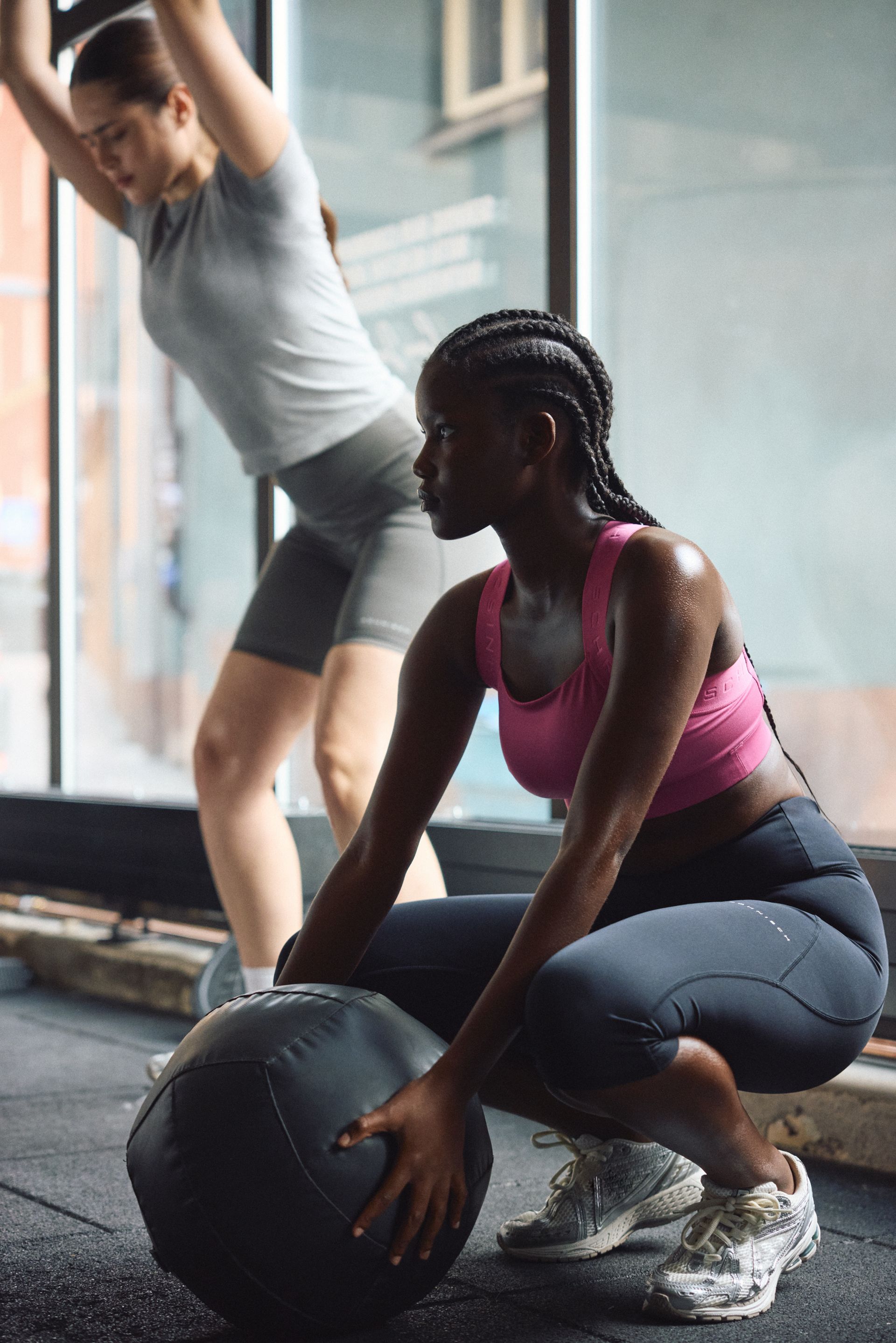 Two women exercising indoors; one squats with a medicine ball, the other lifts her arms. Both wear athletic gear and sneakers.
