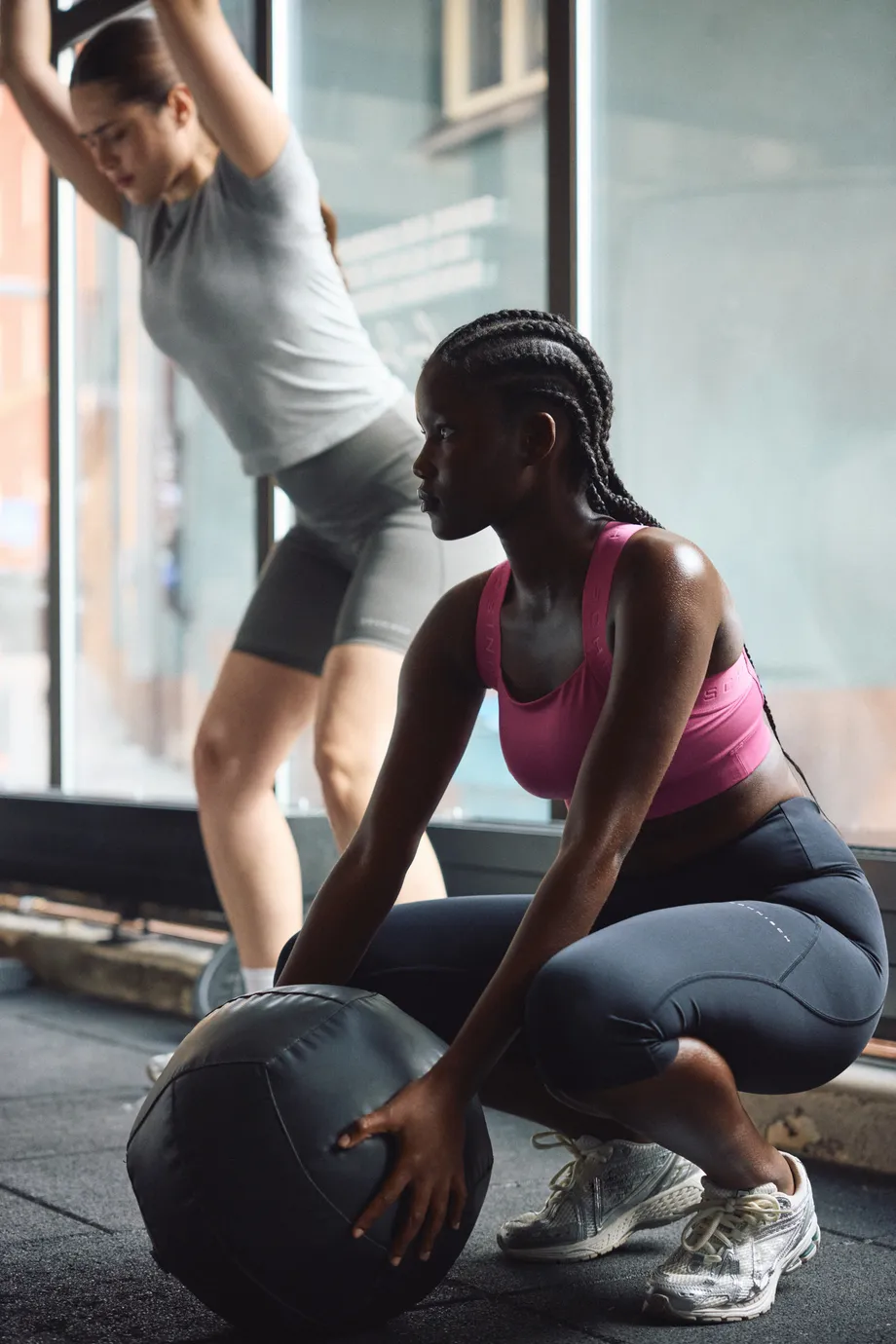 Two women exercising indoors; one squats with a medicine ball, the other lifts her arms. Both wear athletic gear and sneakers.