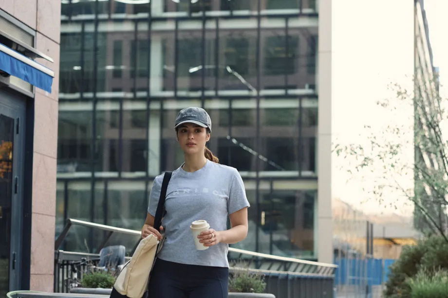 Woman in casual attire walks on a city sidewalk, holding a coffee cup. She wears a cap, gray shirt, and black leggings, with buildings in the background.