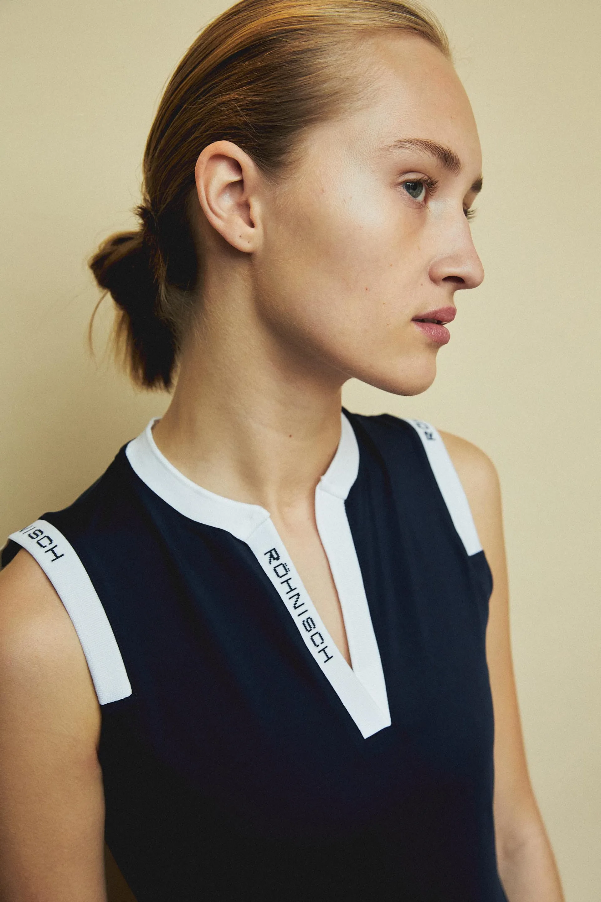 Person in a sleeveless black top with white trim, featuring a side profile against a neutral background. Hair is tied back neatly.