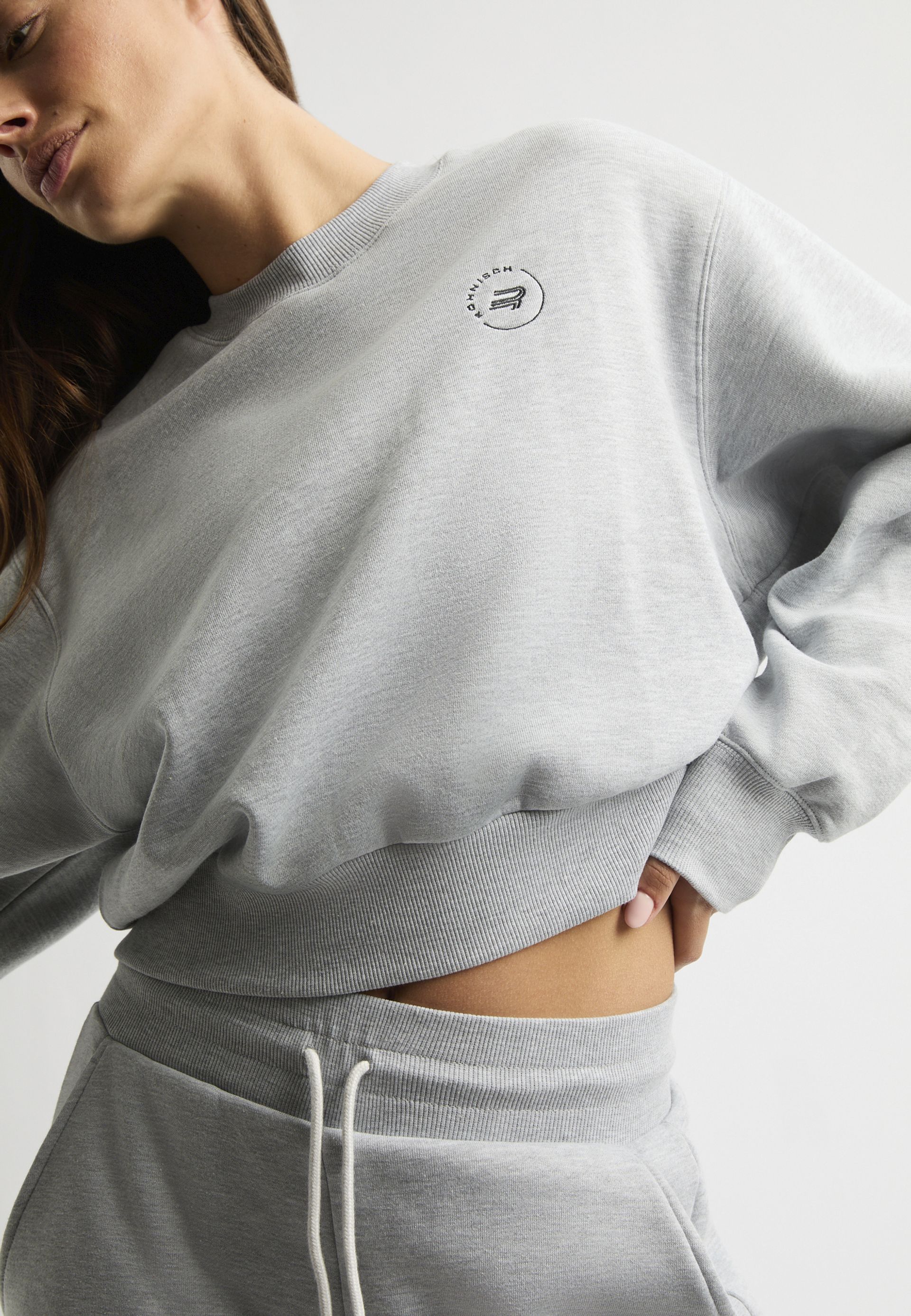 Person wearing a light gray sweatshirt and matching sweatpants, with a small logo on the chest, posing against a plain background.