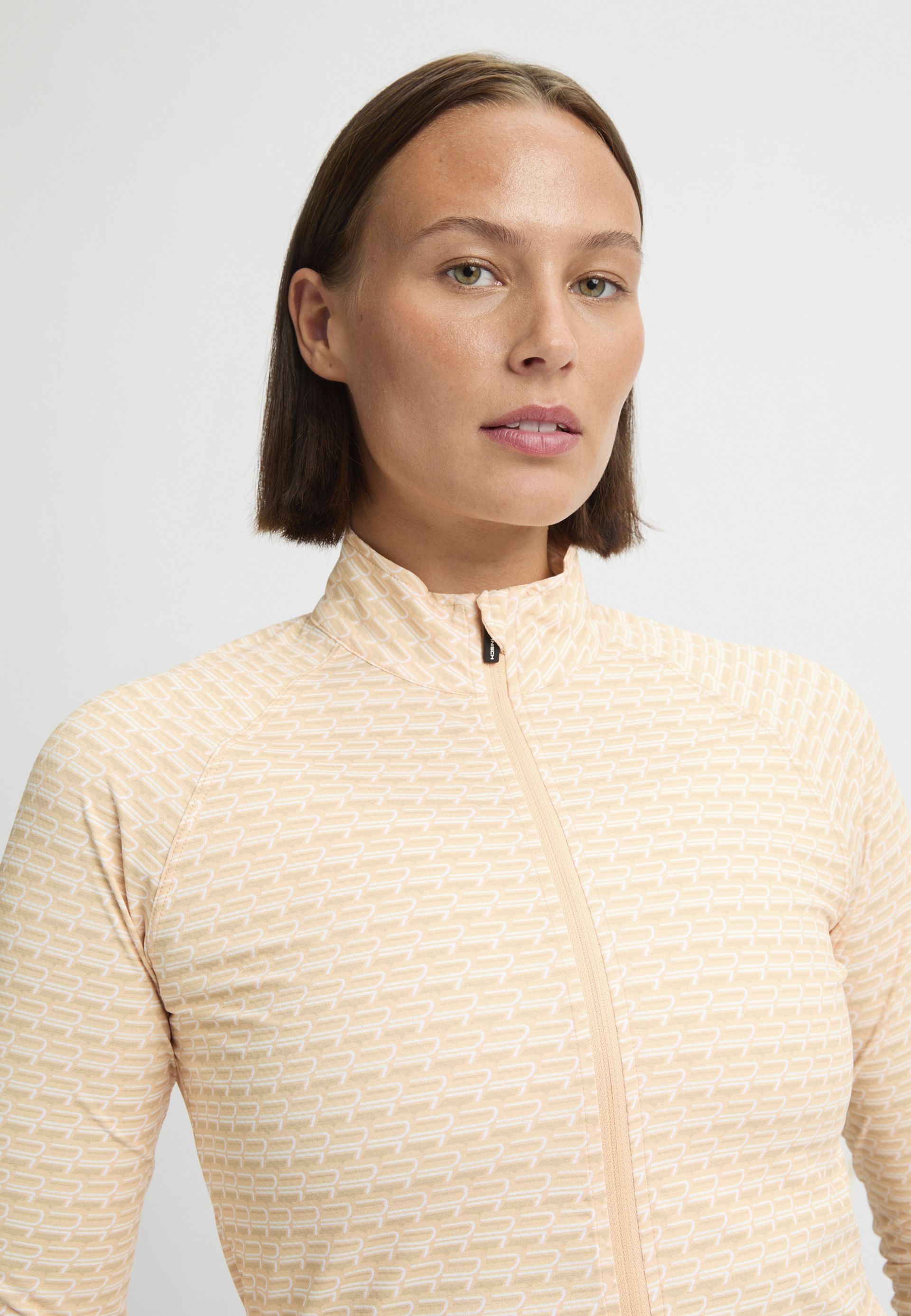 A person wearing a light peach patterned zip-up jacket stands against a plain white background.