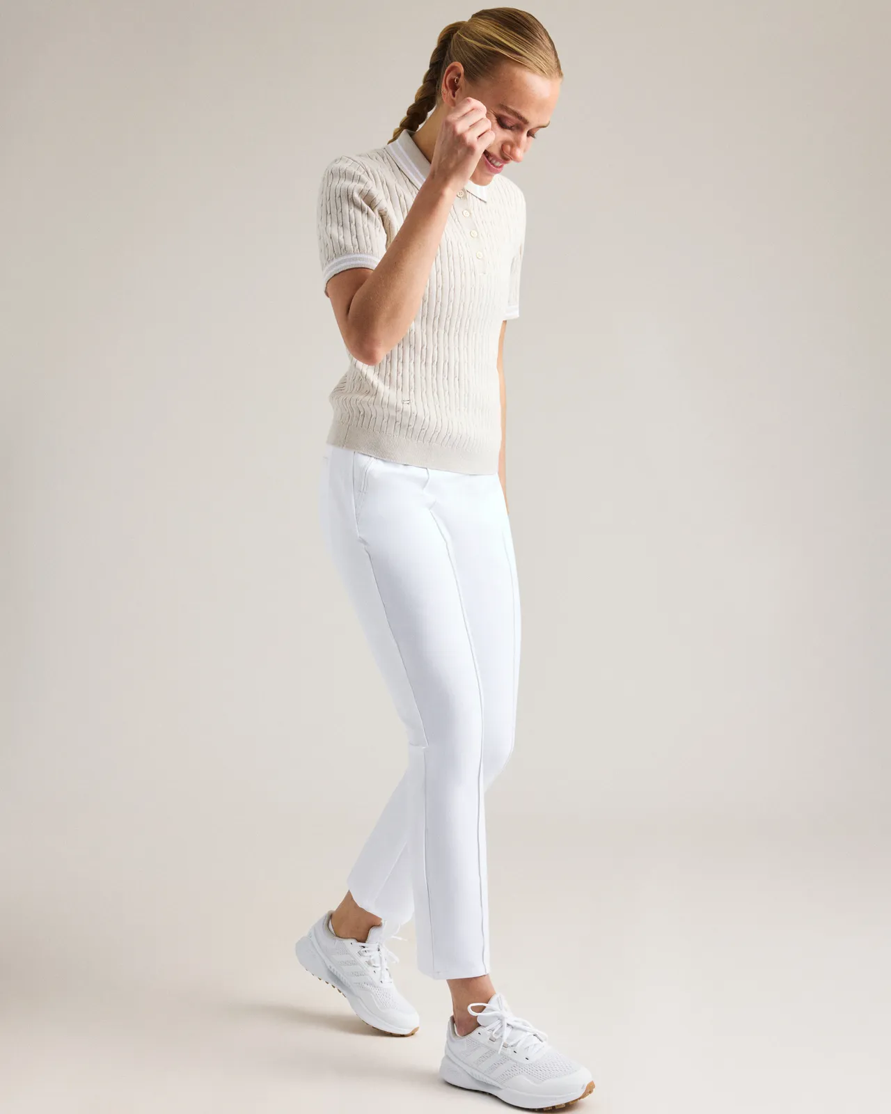 Woman in a cream sweater and white pants, wearing white sneakers, poses against a neutral background.