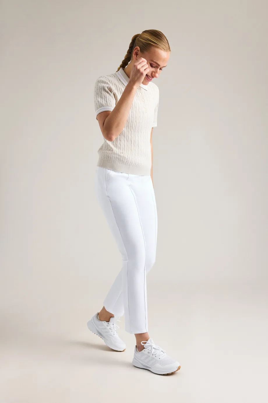 Woman in a cream sweater and white pants, wearing white sneakers, poses against a neutral background.