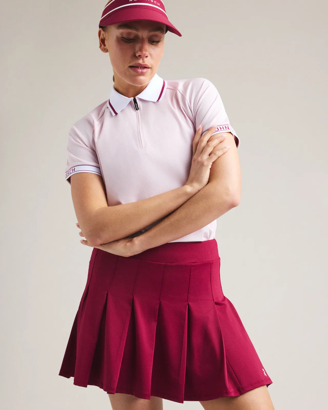 Woman in a pink polo and red pleated skirt with matching visor, arms crossed, against a plain background.