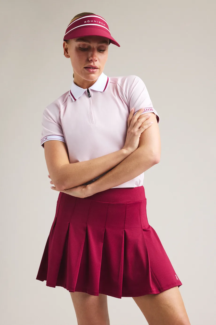Woman in a pink polo and red pleated skirt with matching visor, arms crossed, against a plain background.