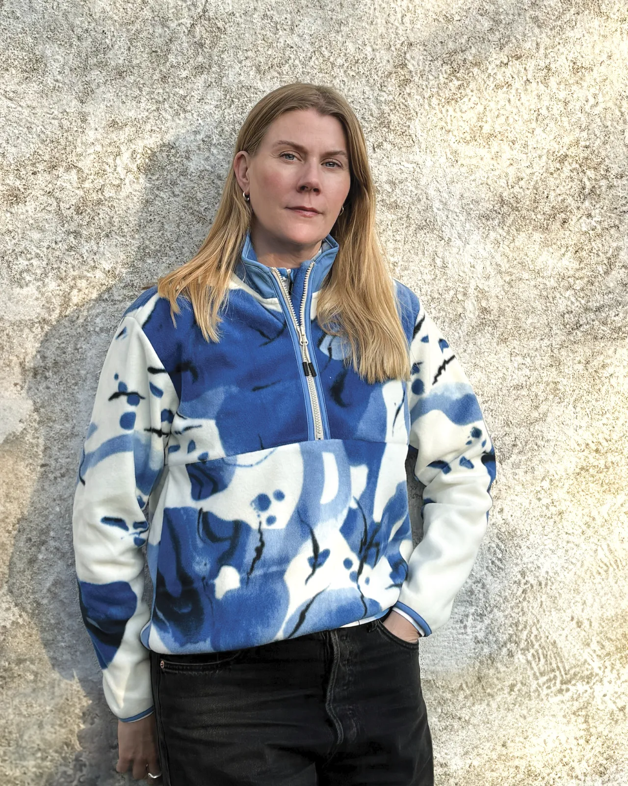 Woman in a blue and white patterned fleece jacket stands against a textured wall, hands in pockets, with long blonde hair and a calm face.
