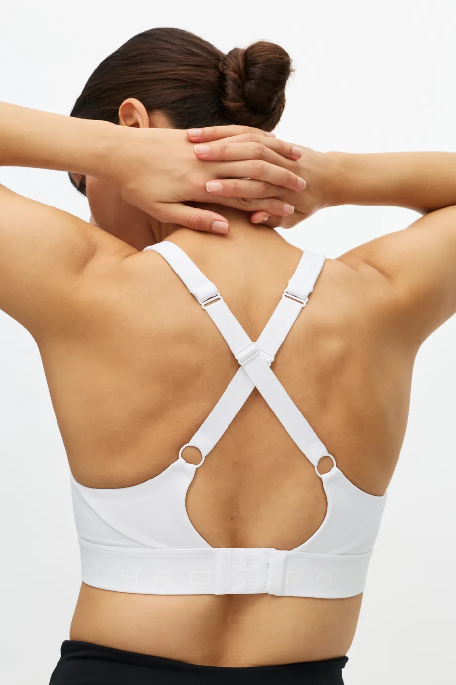 Person with hands on head, wearing a white sports bra with crisscross straps, viewed from the back against a plain background.
