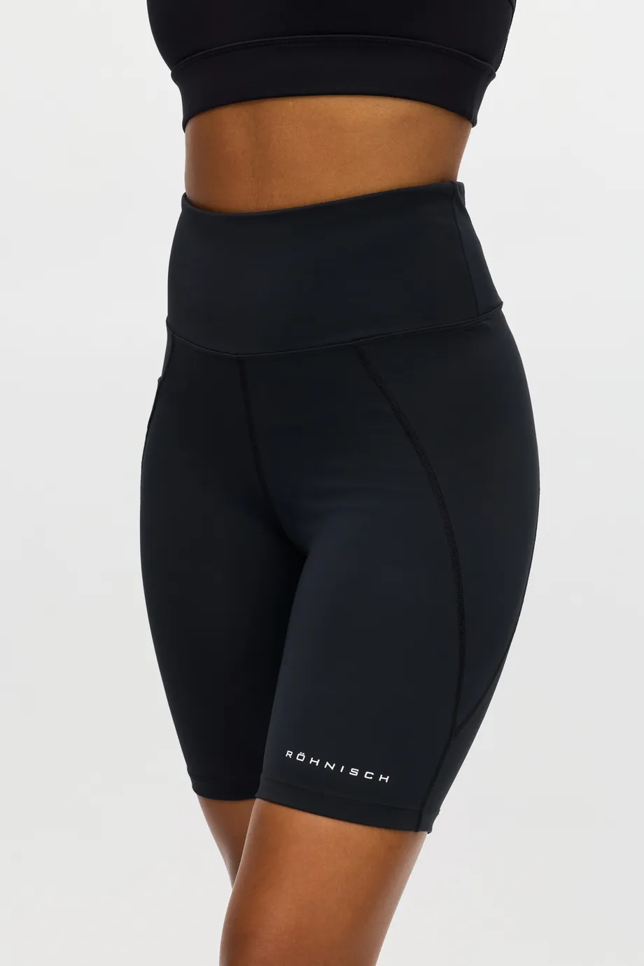 Person wearing high-waisted black athletic shorts and a matching crop top against a plain background. The shorts have branding at the hem.
