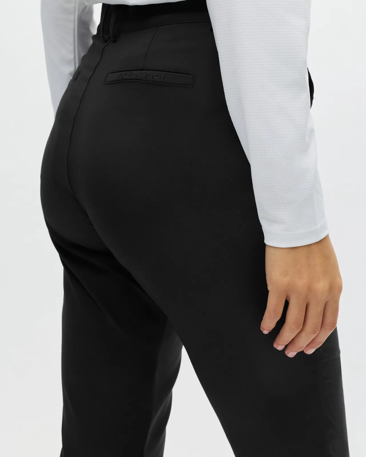 Person in a white long-sleeve shirt and black pants stands with their right hand resting on their hip against a neutral background.