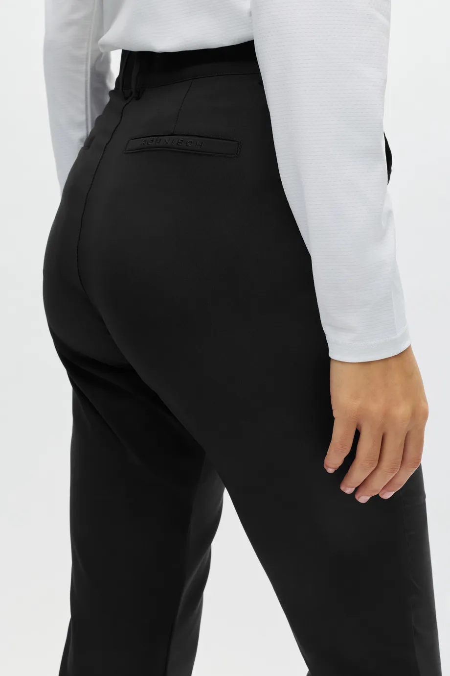 Person in a white long-sleeve shirt and black pants stands with their right hand resting on their hip against a neutral background.