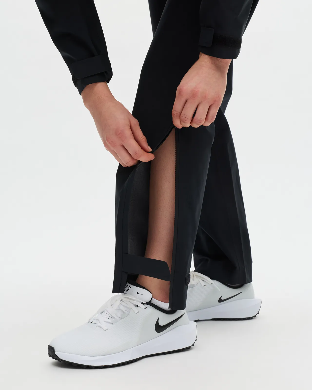Person adjusting zippered ankle of black pants, wearing white Nike sneakers on a white background.