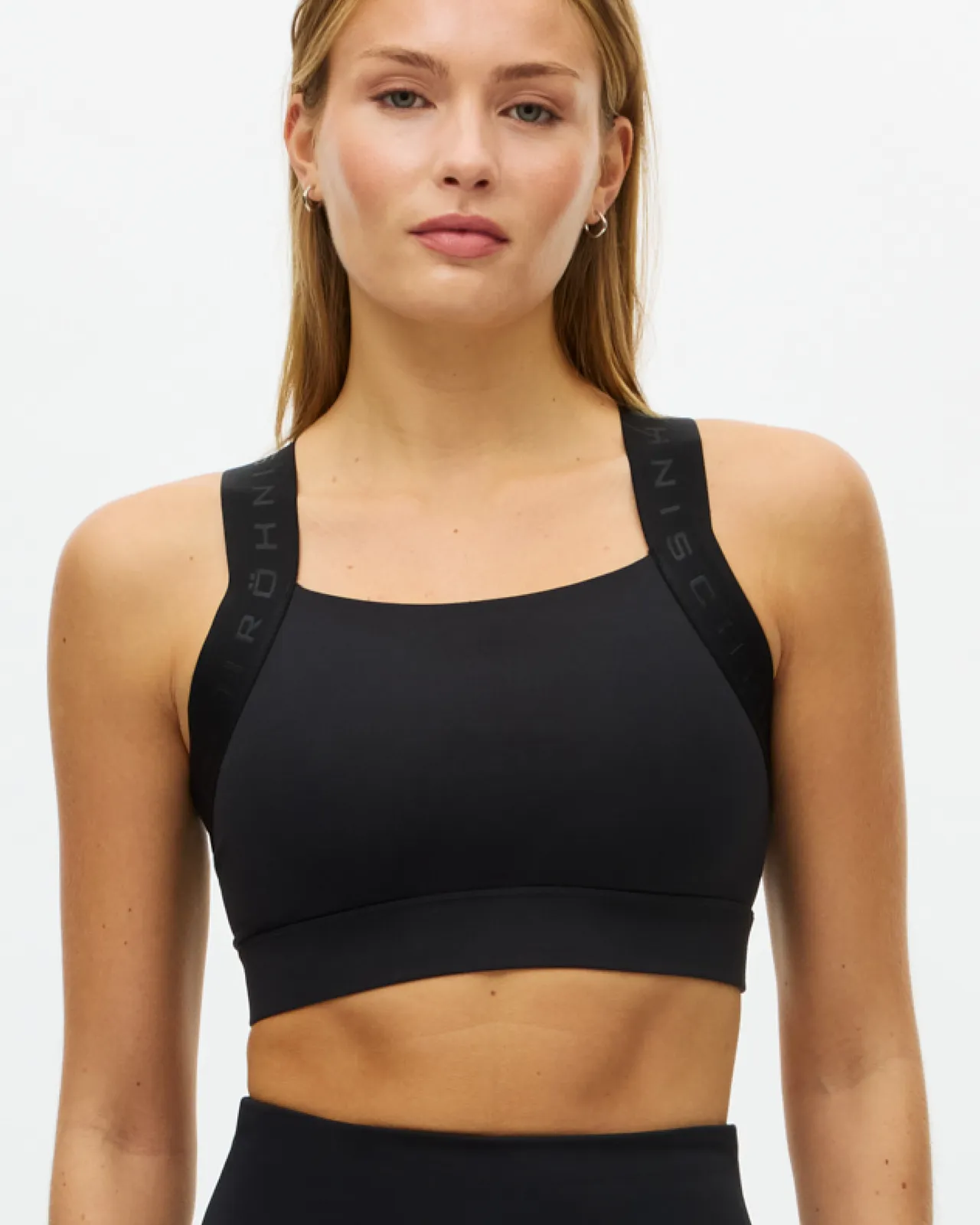 Woman in a black sports bra and leggings stands against a plain white background, looking confidently at the camera.