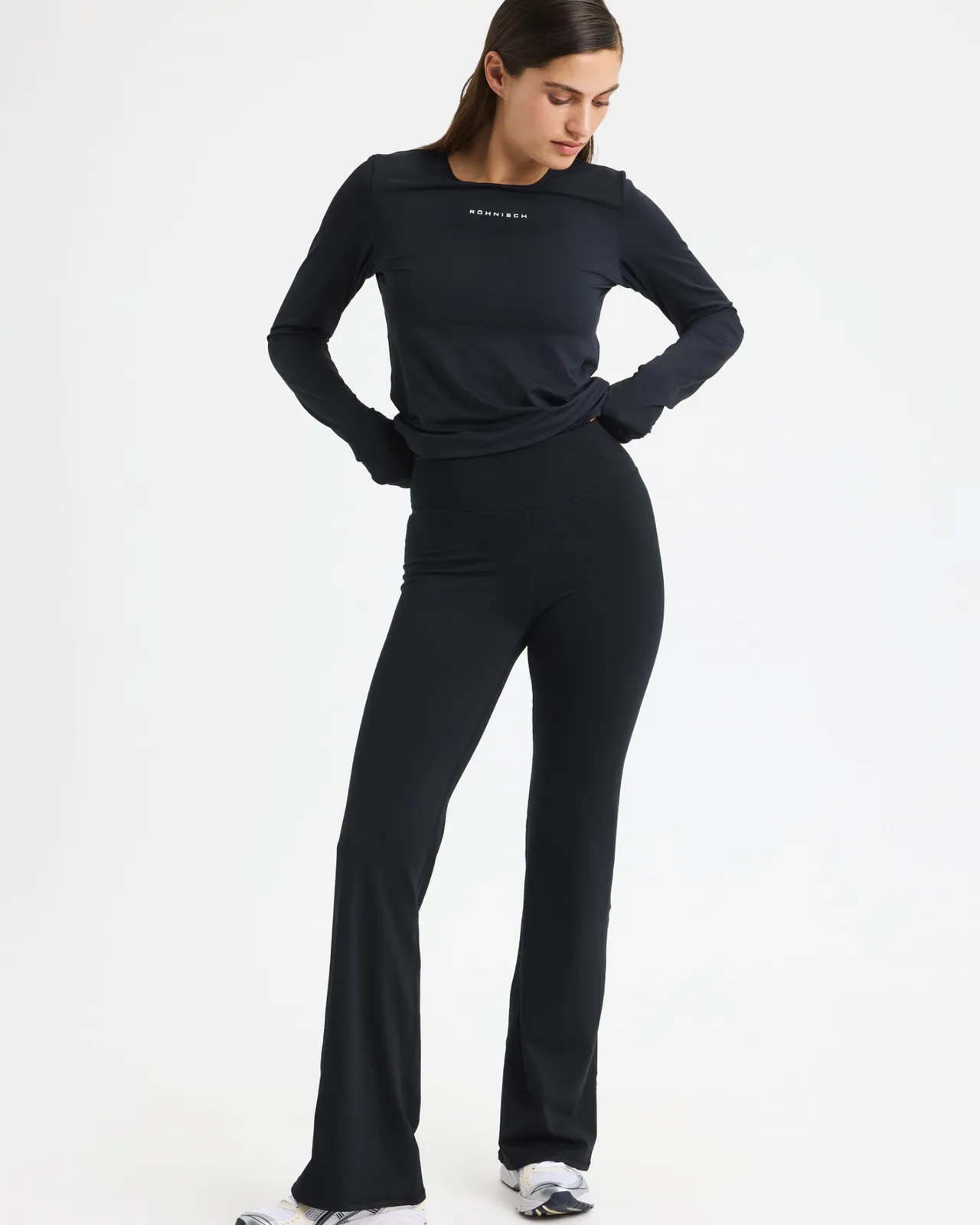 Woman in a black long-sleeve top and flared pants stands with hands behind her back, wearing white sneakers against a plain background.