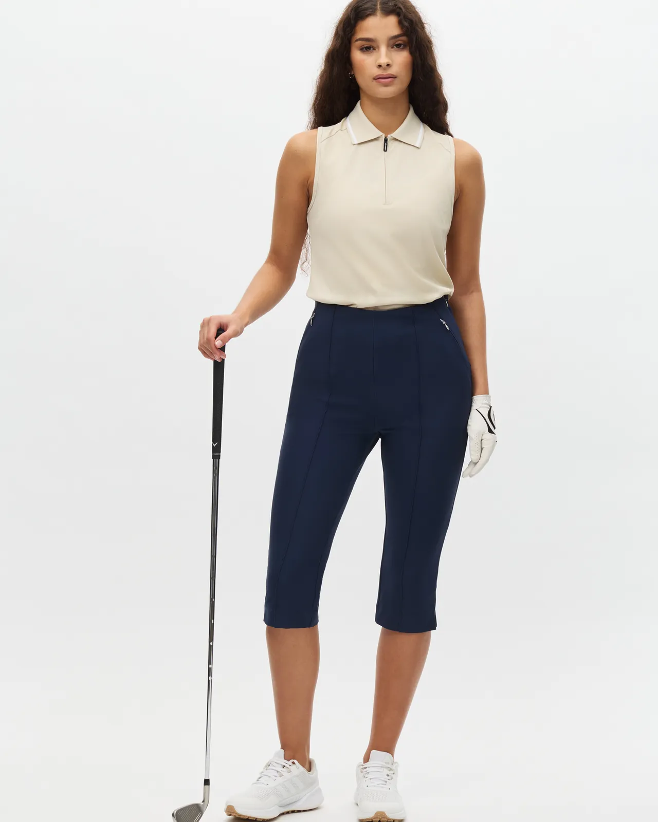 Woman with long hair in beige sleeveless polo and navy pants holds a golf club, wearing a white glove and sneakers, standing on a white background.