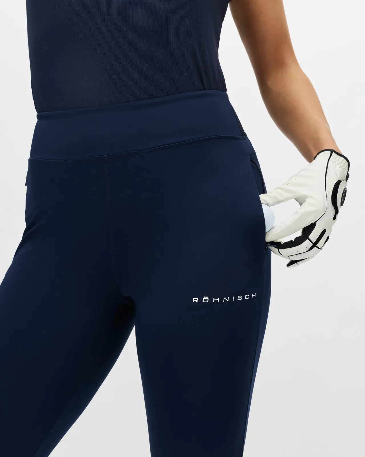 Person wearing navy athletic leggings with a logo, holding a pair of sports gloves in one hand against a white background.