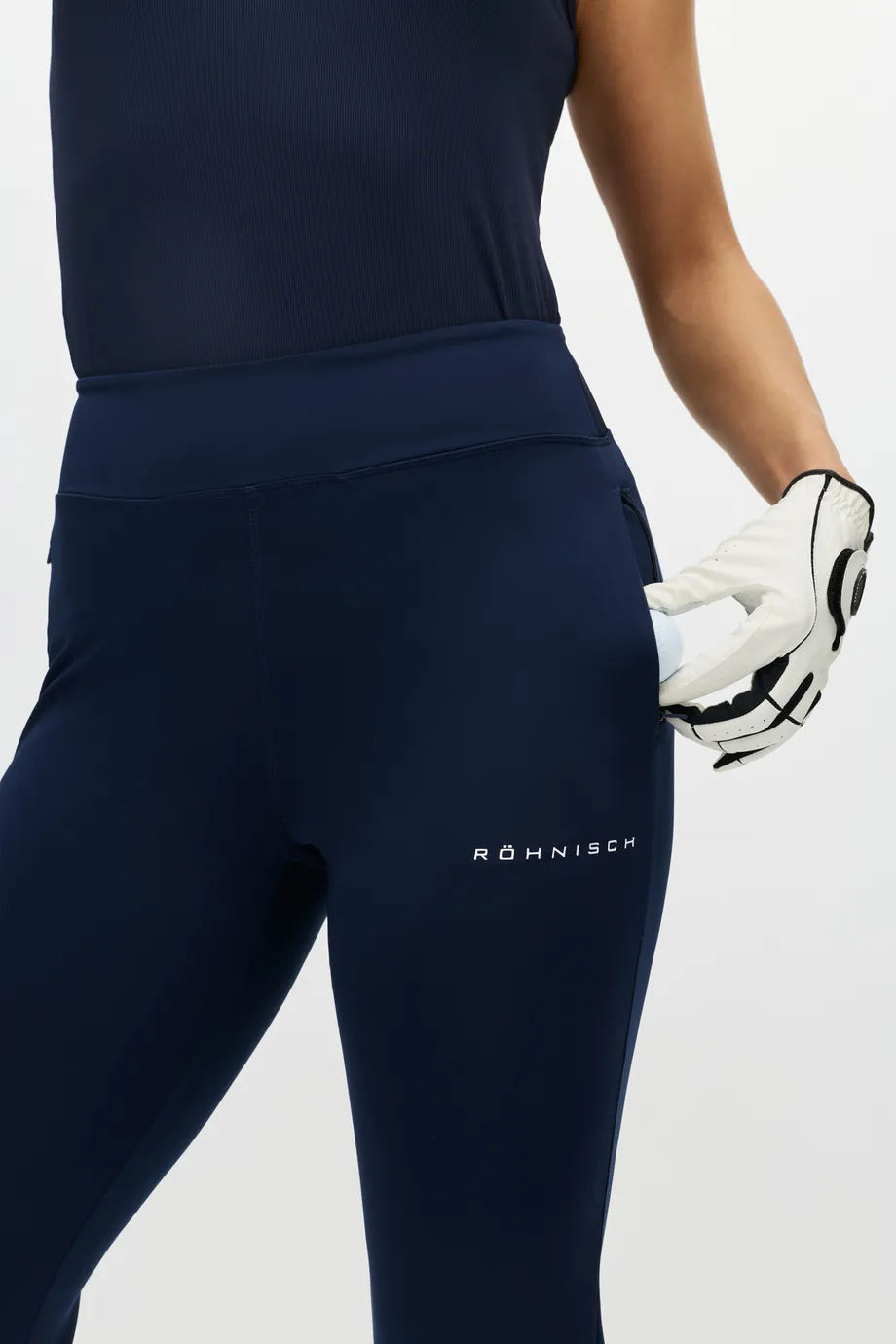 Person wearing navy athletic leggings with a logo, holding a pair of sports gloves in one hand against a white background.