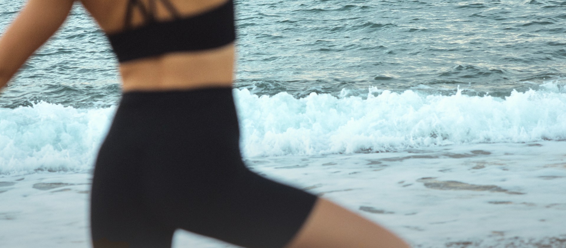 A person in black athletic wear balances on one leg at a pebbled beach, with ocean waves in the background.